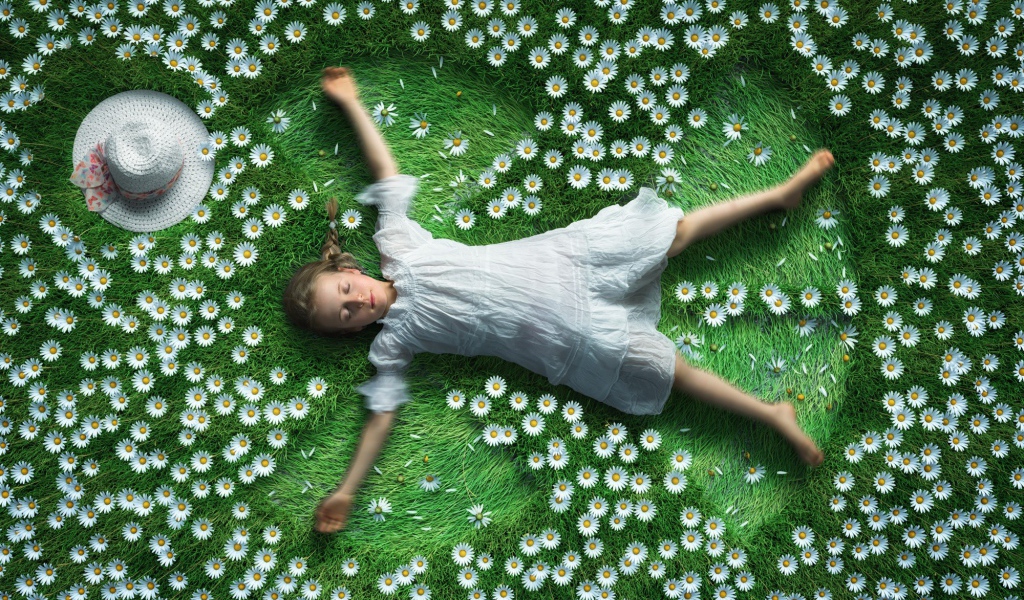 Маленькая девочка в белом платье лежит на зеленой траве с ромашками