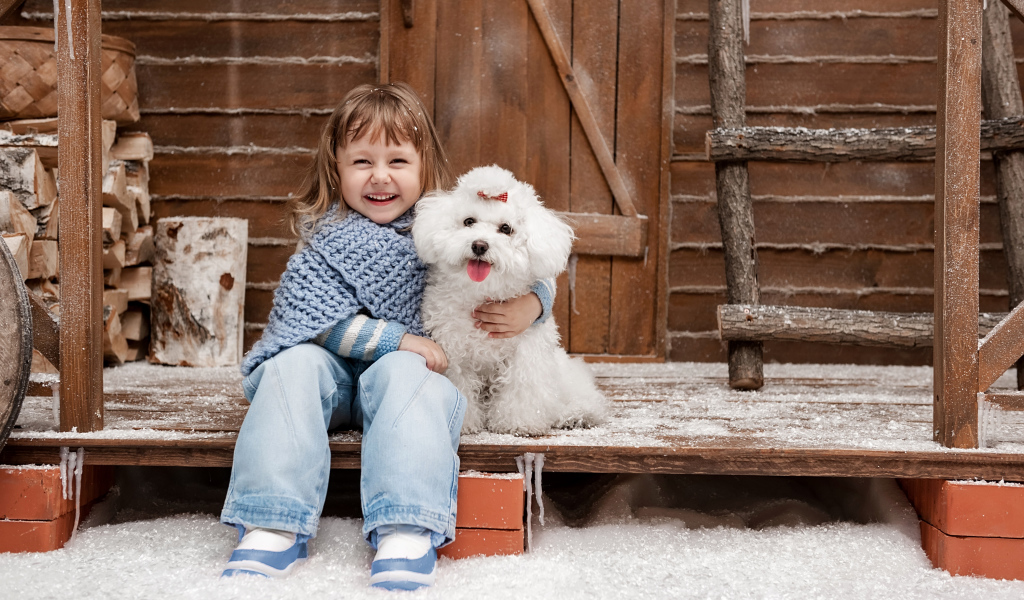 Маленькая улыбающаяся девочка сидит с белым пуделем на крыльце дома зимой