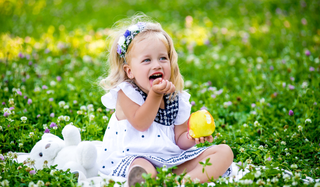 Маленькая улыбающаяся девочка с яблоком сидит на зеленой траве