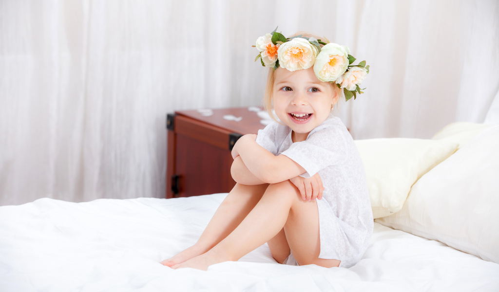 Улыбающаяся девочка с венком на голове сидит на кровати