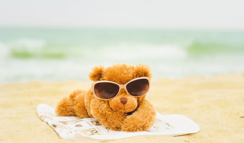 Плюшевый мишка в солнечных очках лежит на пляже