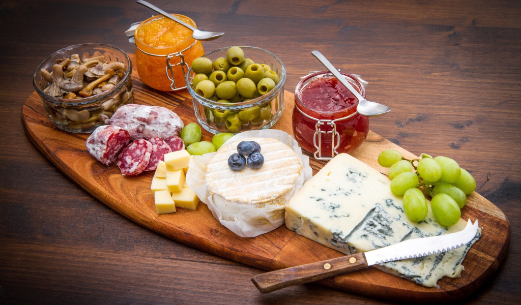 Сыр, грибы, маслины, колбаса, виноград и джем на разделочной доске 