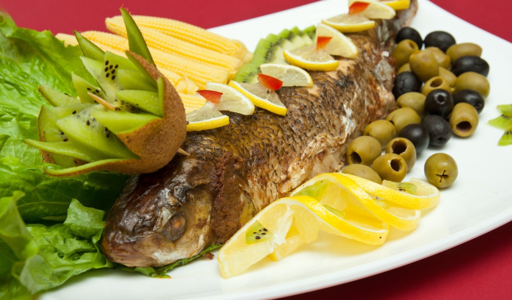 Запеченная рыба с оливками, лимоном, киви, кукурузой и листьями салата на белой тарелке