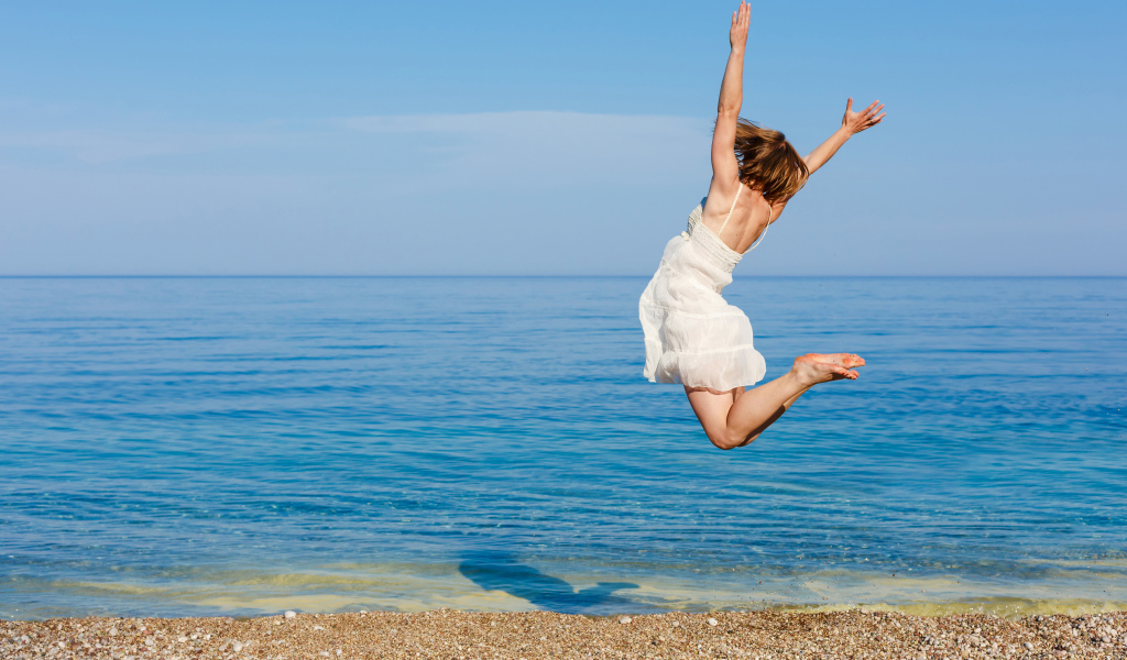 A young girl in a white dress is making a jump on the sand by the sea