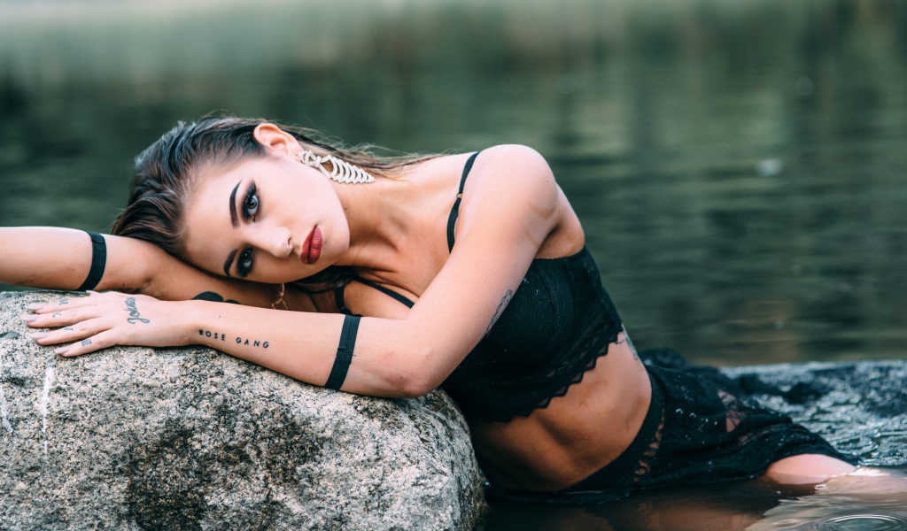 Молодая девушка с татуировками сидит у камня в воде