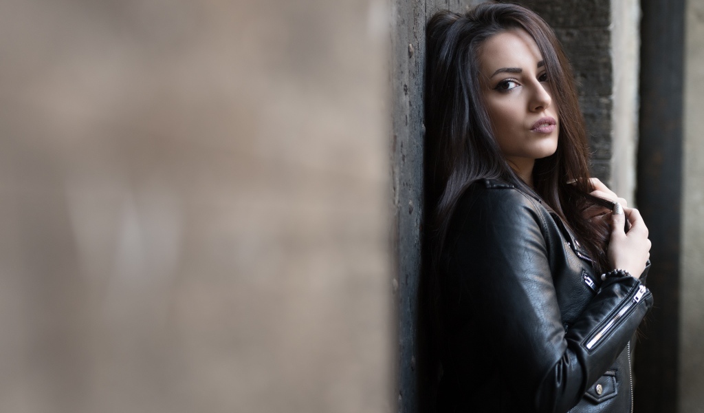 Красивая  брюнетка в кожаной куртке стоит у стены