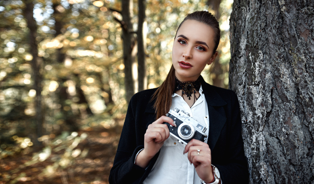 Красивая девушка с фотоаппаратом в руках стоит у дерева