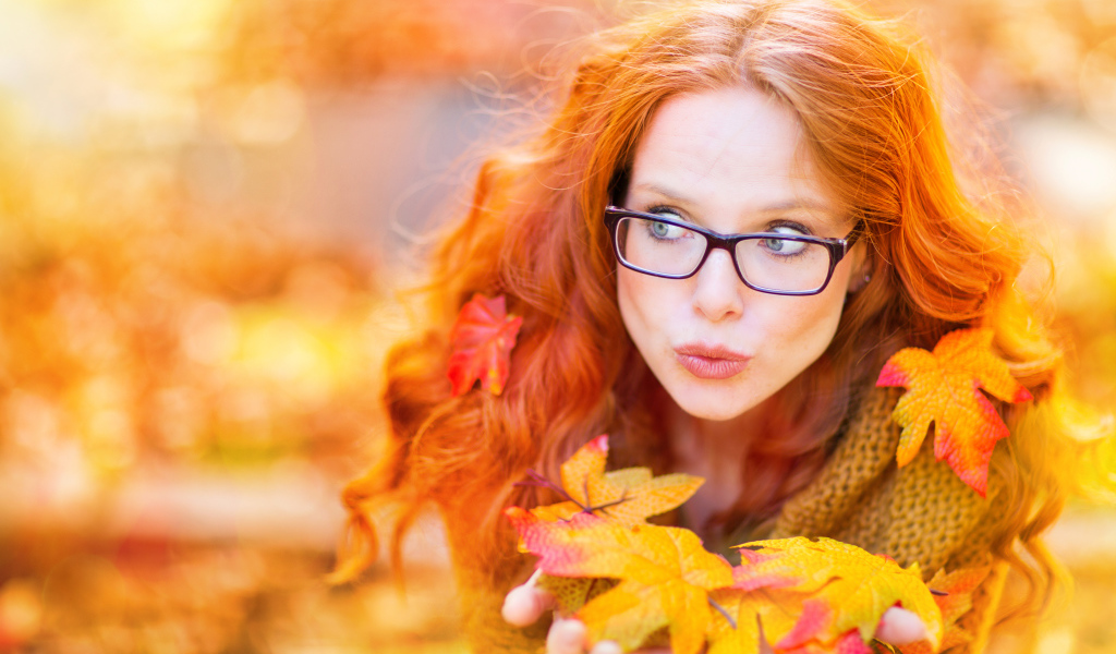 Рыжеволосая девушка в очках держит в руках опавшие листья 