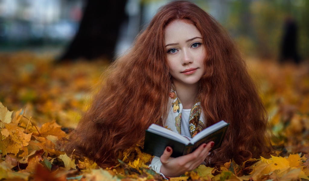Рыжеволосая девушка с книгой лежит на опавших листьях осенью 