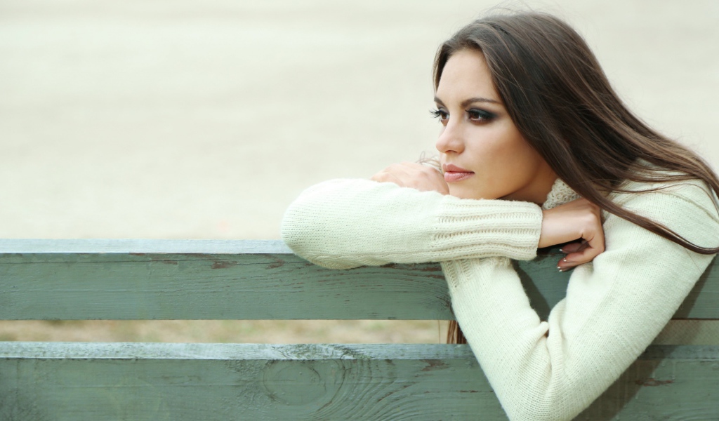 Задумчивая девушка брюнетка в белом свитере сидит на лавке