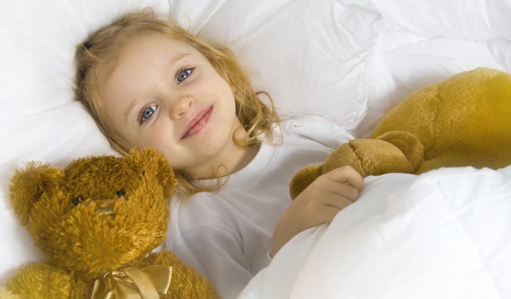 Милая голубоглазая девочка с мягкими игрушками лежит под одеялом