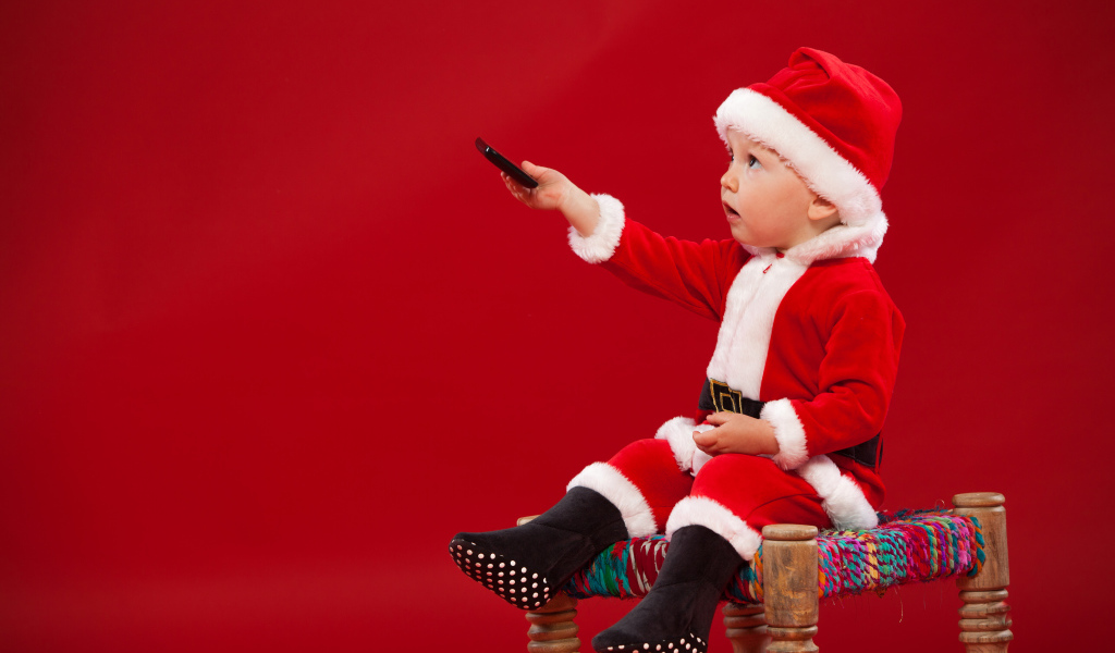 Маленький мальчик в костюме Санта Клауса сидит на стуле на красном фоне