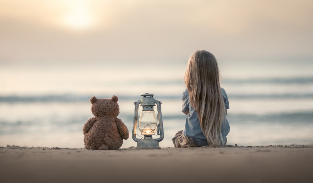 Маленькая девочка сидит на песке с плюшевым мишкой 