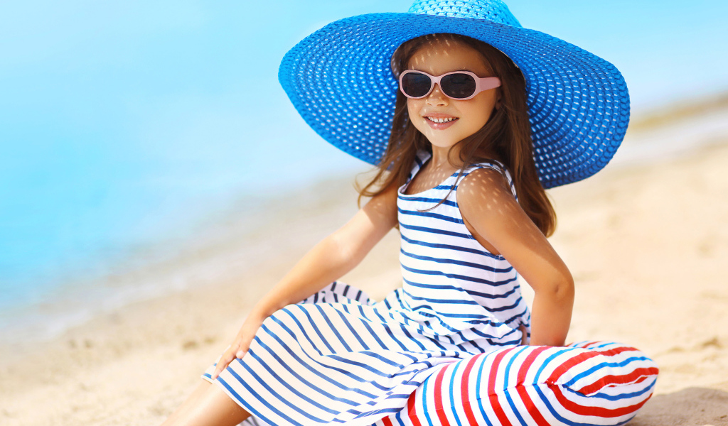 Маленькая девочка сидит на подушке на пляже в большой синей шляпе