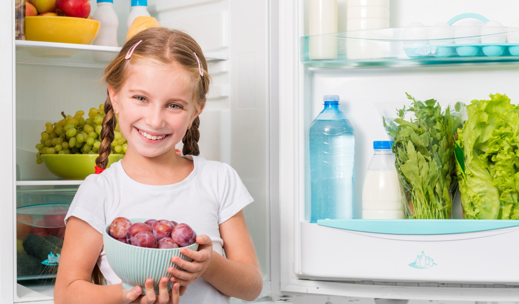 Маленькая девочка с миской со сливами в руках у холодильника 