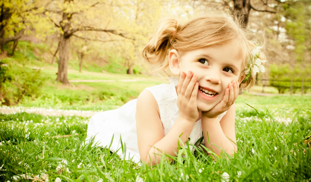 Улыбающаяся девочка в белом платье лежит на зеленой траве