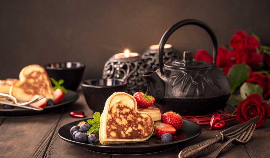Оладьи в форме сердца с ягодами черники и клубники на столе с чаем и свечами