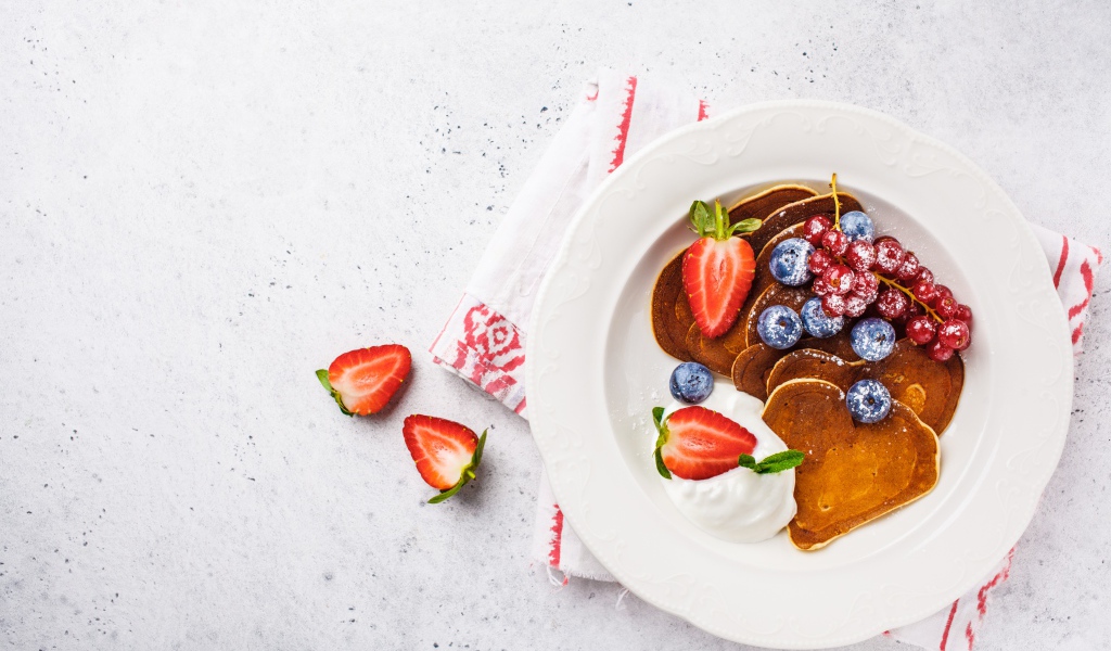 Оладьи со сметаной в тарелке с ягодами черники, клубники и красной смородины