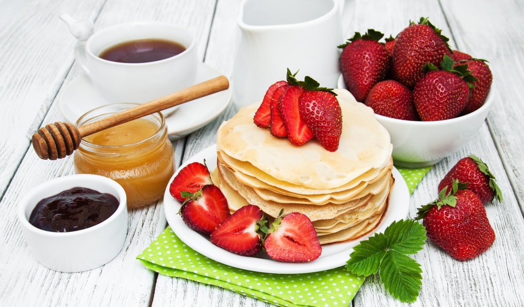 Тонкие блины на столе с ягодами клубники, медом, чаем и джемом