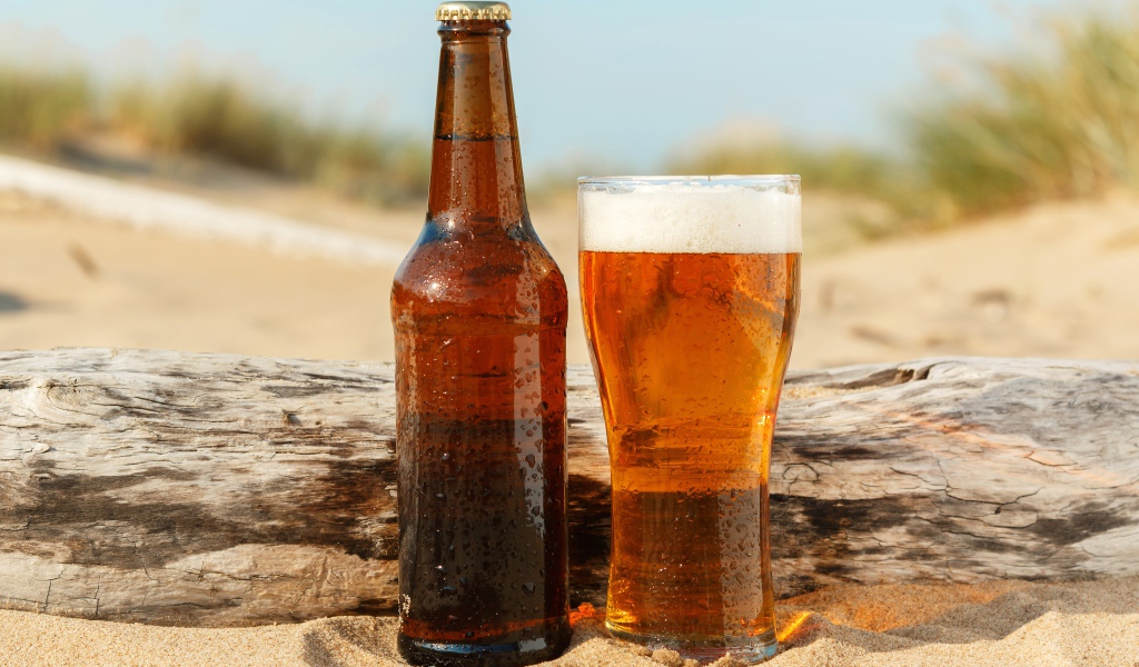Бутылка пива и бокал стоят на песке 