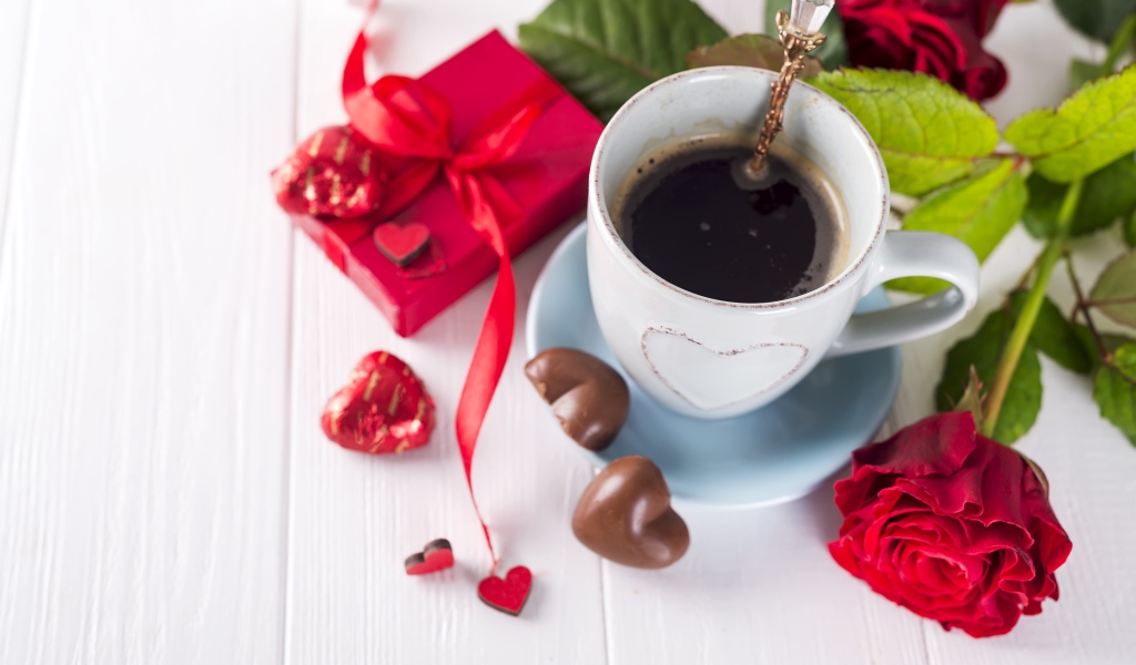 Чашка кофе на столе с шоколадными конфетами, подарком и красной розой