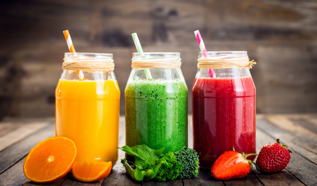 Три банки смузи с трубочкой на столе с апельсином, брокколи и клубникой