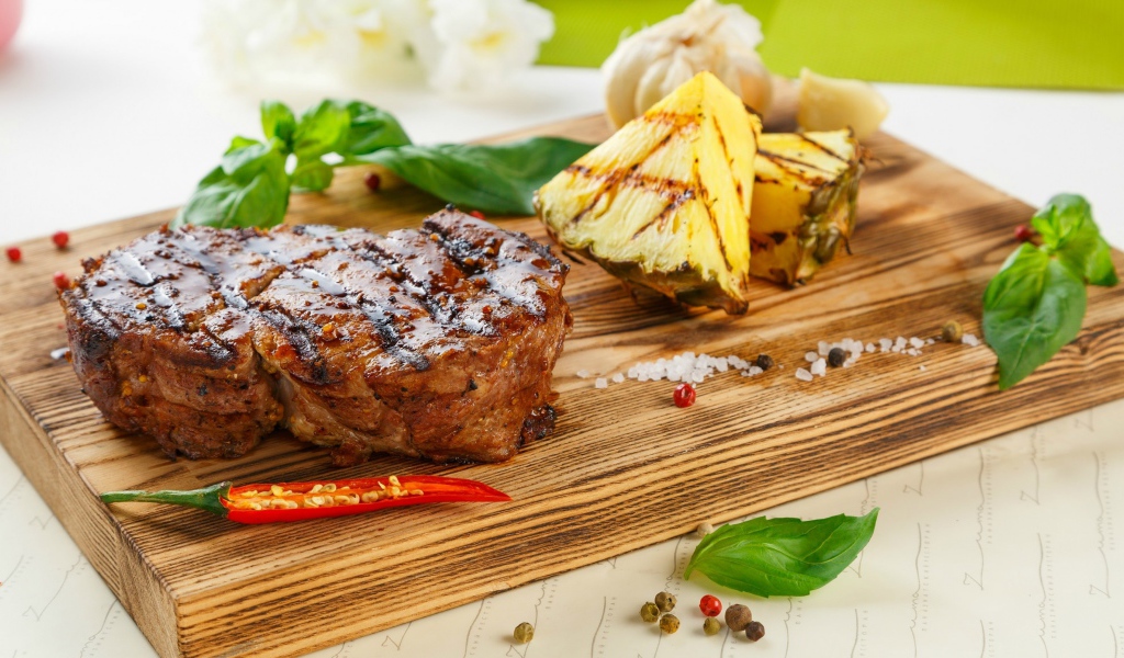 A piece of grilled meat on a wooden board with pineapple slices and basil