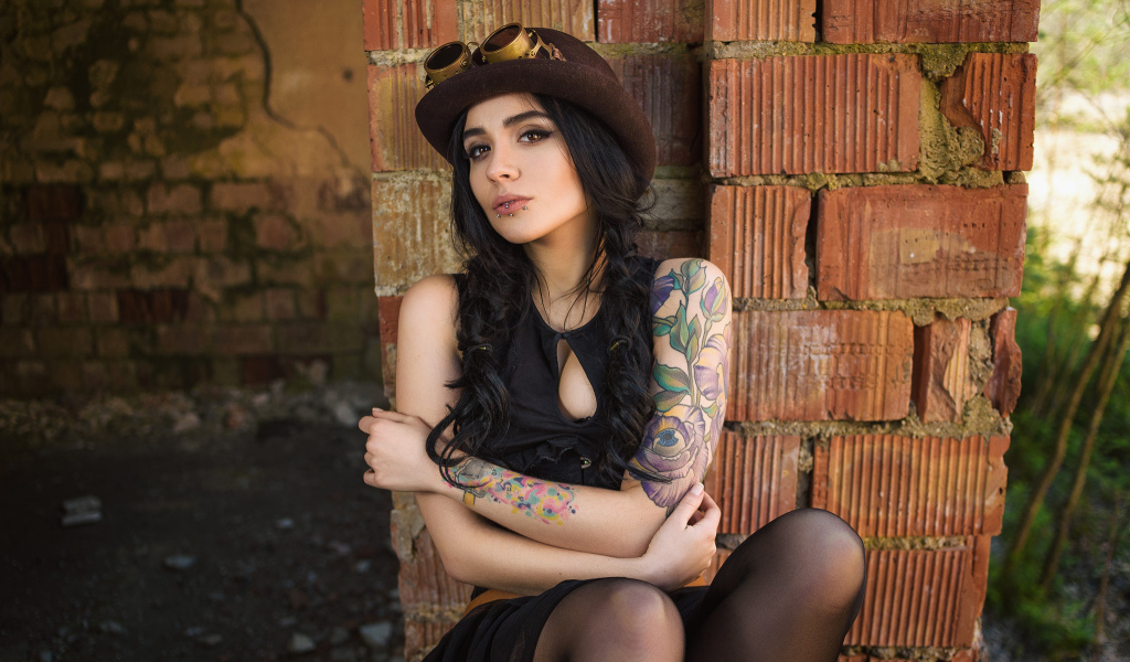 Девушка в шляпе с татуировками на руке сидит у стены