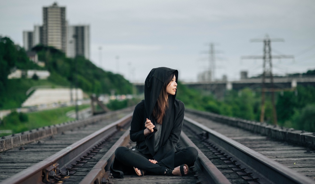 Девушка азиатка в черном балахоне сидит на рельсах 