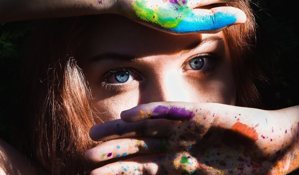 Красивая голубоглазая девушка с руками в разноцветной краске у лица