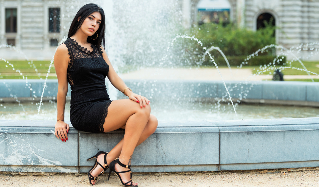 Красивая брюнетка в черном платье сидит у фонтана