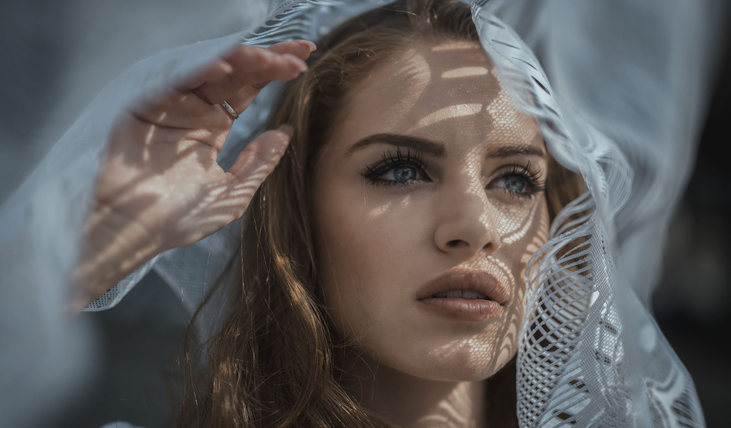 Голубоглазая девушка смотрит в окно с тюлью