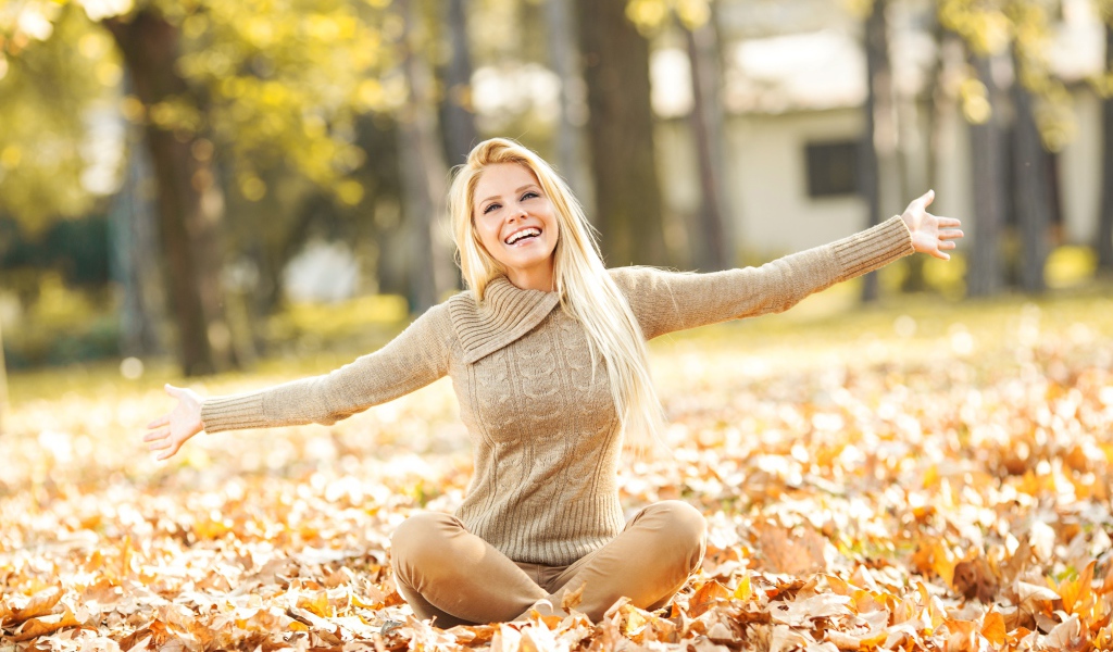 Веселая девушка сидит на опавшей листве осенью 