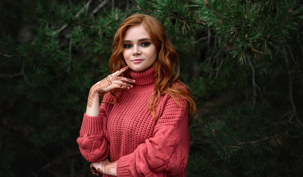 Рыжеволосая девушка в розовом свитере стоит у ели