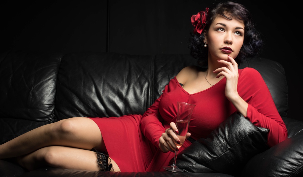 Девушка в красном платье с бокалом в руке лежит на кожаном диване