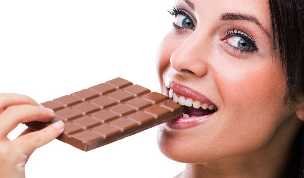 Молодая девушка кусает молочную шоколадку на белом фоне