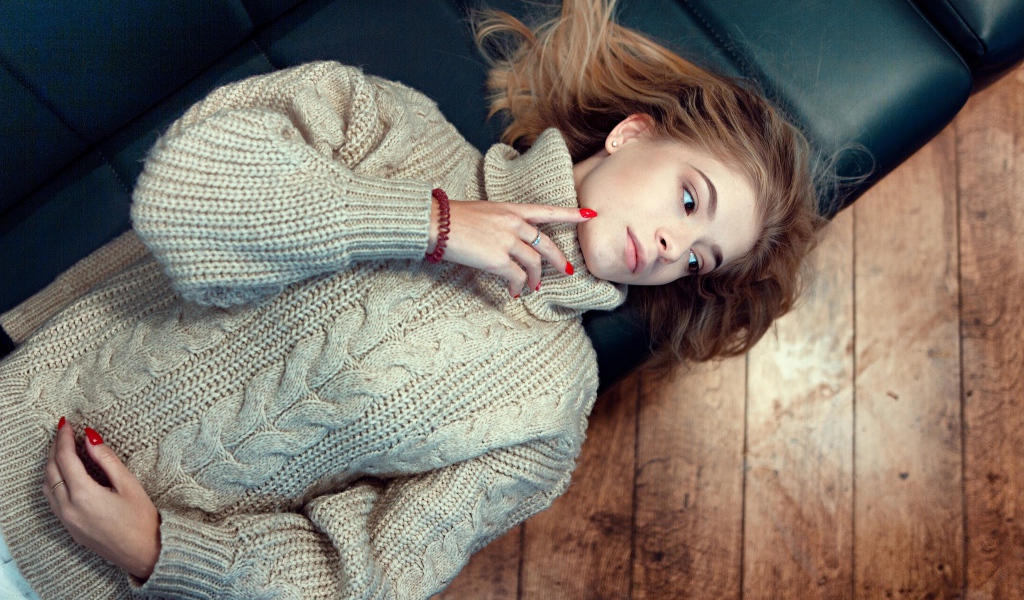 Молодая девушка в вязаном свитере лежит на черном кожаном диване