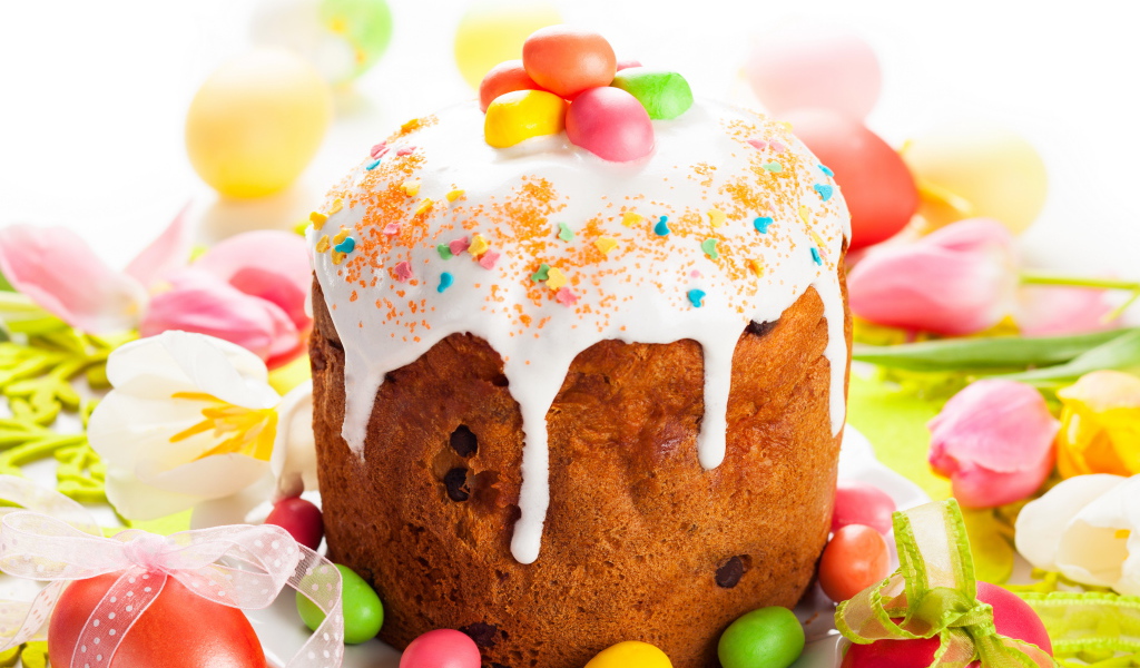 Красивый пасхальный кулич с белой глазурью и конфетами на столе с яйцами