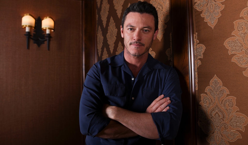 Актер Люк Эванс в рубашке стоит у стены