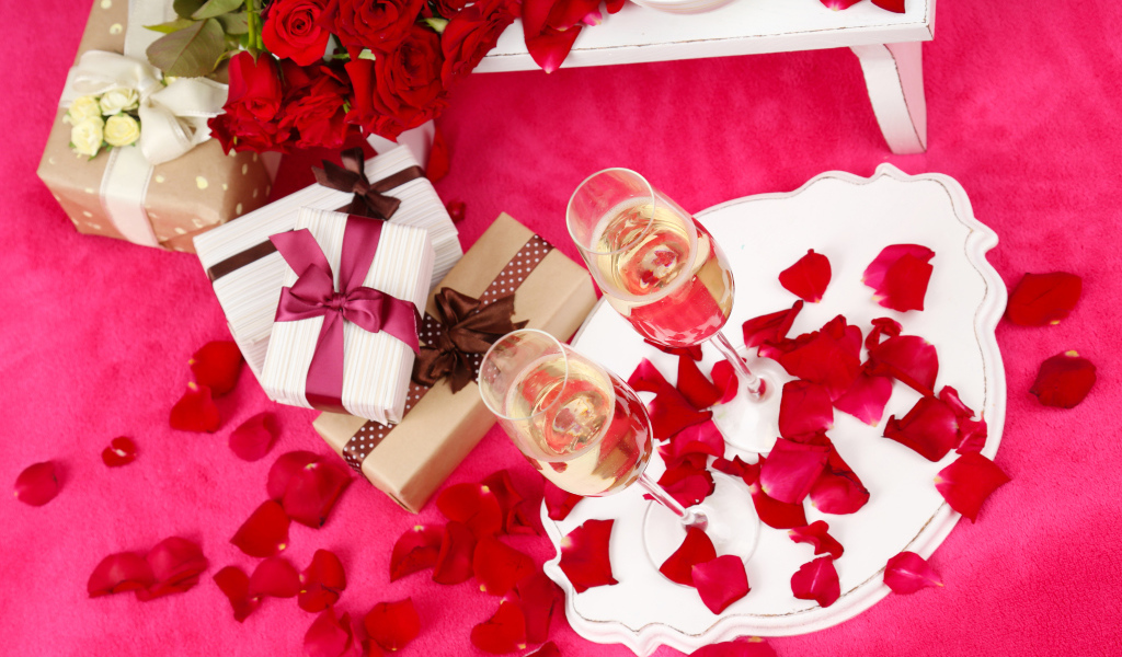 Два фужера с шампанским на красном покрывале с лепестками, подарками и букетом роз для любимой