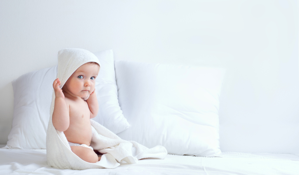 Маленький мальчик сидит в белом покрывале на кровати с подушками