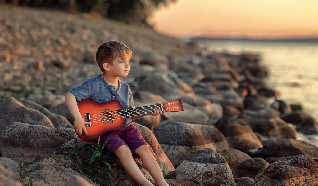 Маленький мальчик сидит с гитарой на камнях у моря 