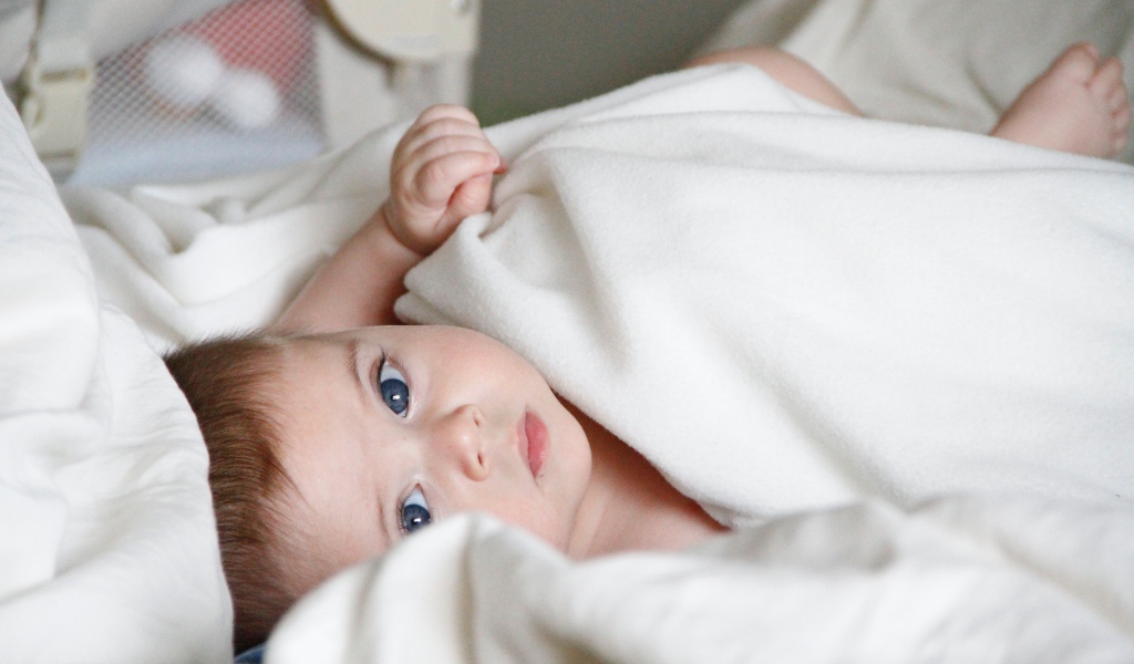 Маленький грудной голубоглазый ребенок лежит под белым покрывалом 