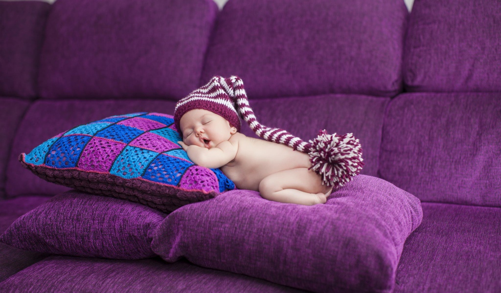 Маленький ребенок в большой шапке спит на фиолетовых подушках