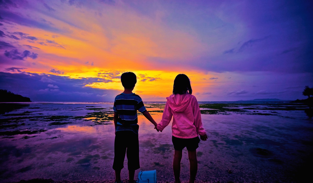 Маленькие мальчик и девочка стоят у воды на фоне заката 