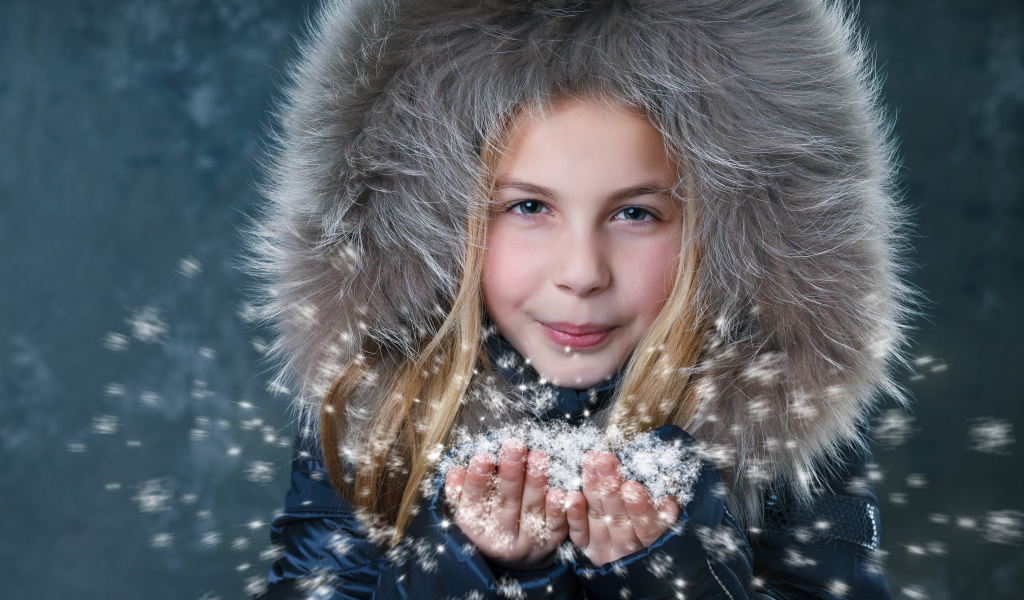 Маленькая девочка в капюшоне сдувает снежинки с ладоней