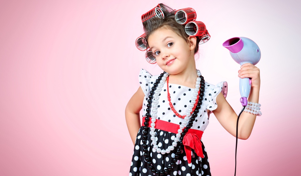 Маленькая девочка с бигудями на голове с феном в руке на розовом фоне