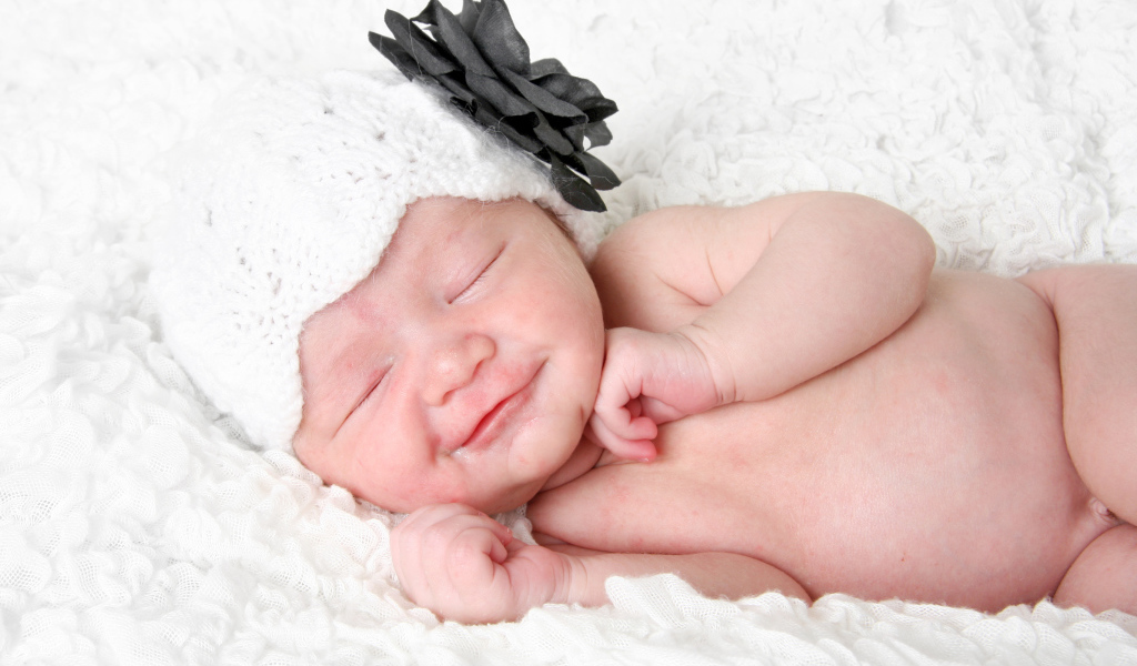 Улыбающийся спящий ребенок в белой вязаной шапке с цветком