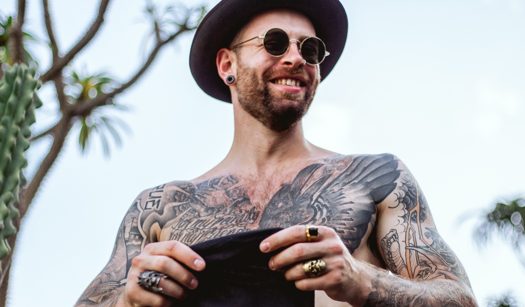 Мужчина с татуировками на теле в черной шляпе и очках
