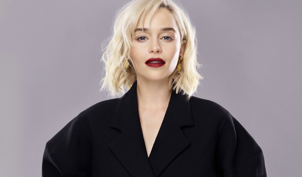 Яркая блондинка, актриса Эмилия Кларк в черном пальто на сером фоне